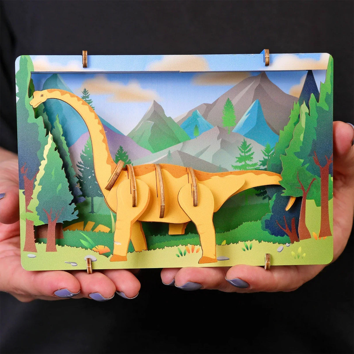 Hände halten ein 3D Holzmodell eines Dinosauriers in einer Wald und Berglandschaft