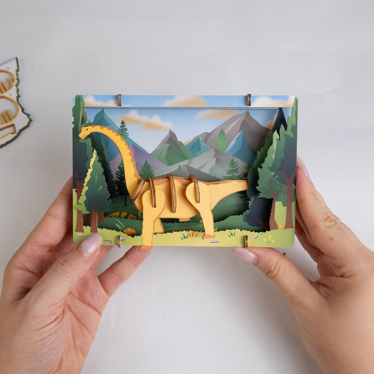 Hände halten ein mehrschichtiges 3D-Papier-Diorama mit einem Dinosaurier vor einer Berglandschaft