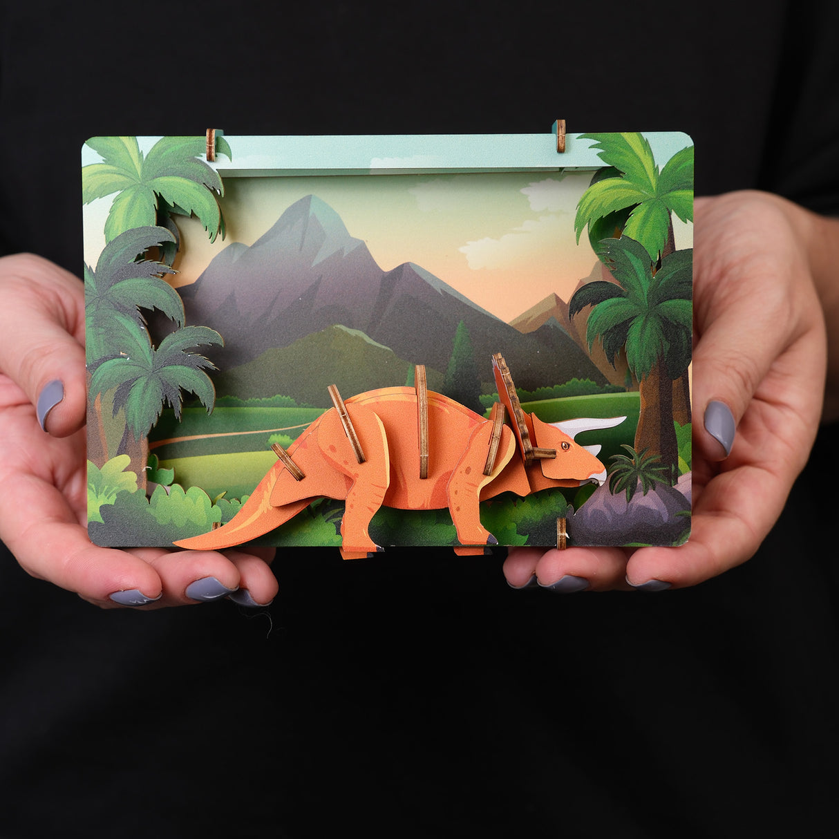 Haende halten ein 3D Holzmodell eines Triceratops in einer Dschungellandschaft