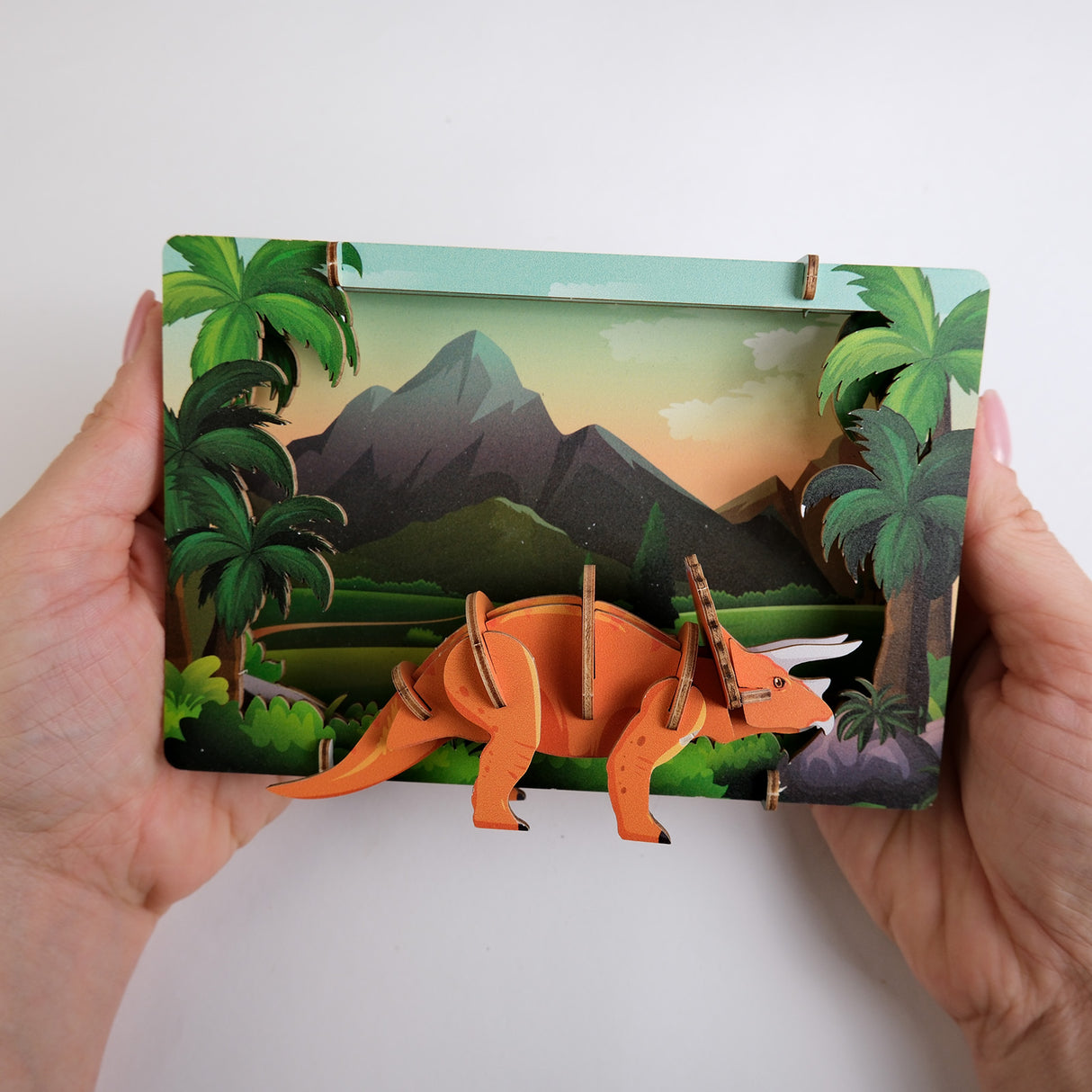 Hände halten ein 3D Holzmodell eines Triceratops vor einer illustrierten Berglandschaft