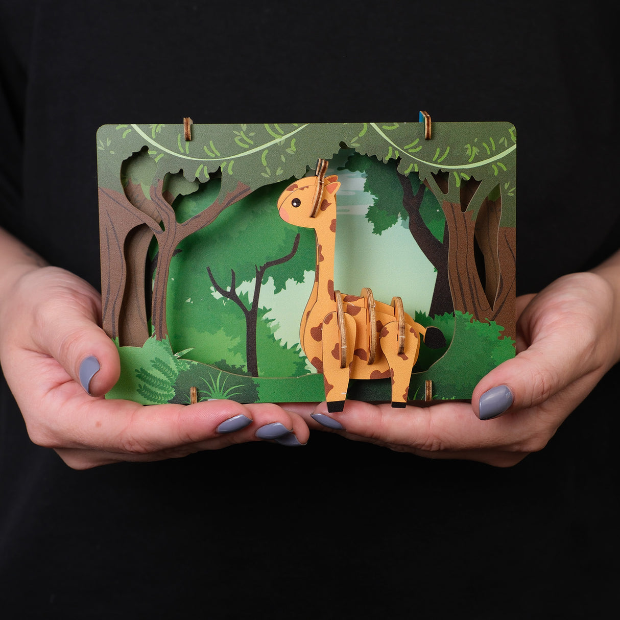 Haende halten ein dreidimensionales Giraffenmodell aus Pappe vor einer Waldkulisse