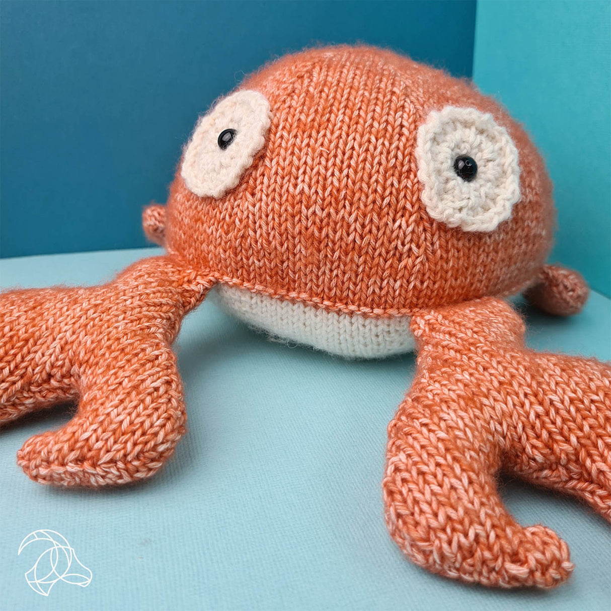 Gestricktes Stofftier in Form einer orangefarbenen Krabbe mit hellen Augen auf blauem Hintergrund