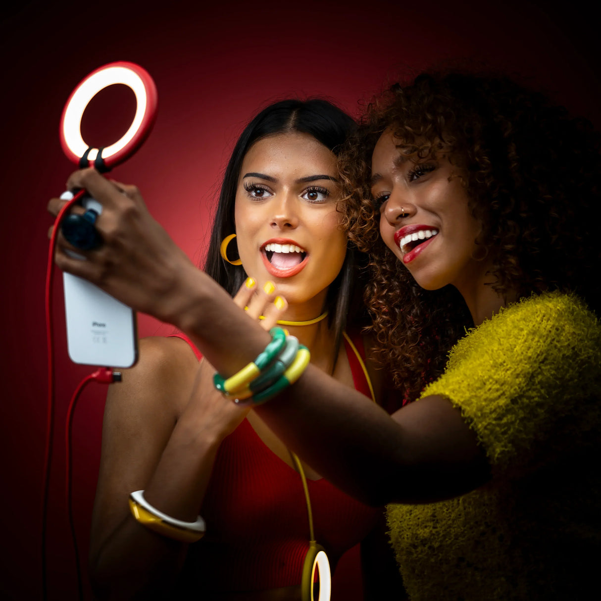Zwei Frauen machen ein Selfie mit einem Smartphone und einem Ringlicht vor rotem Hintergrund