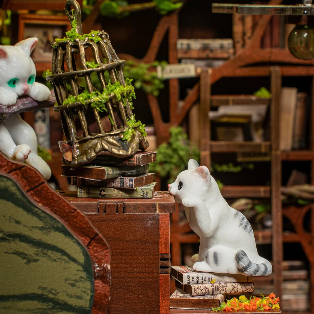 Zwei Katzenfiguren in einem Bücherregal, eine sitzt auf Büchern, die andere schaut auf einen umgestürzten Käfig