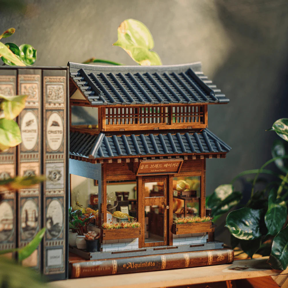Miniatur-Bäckerei-Diorama mit Holzwänden und blauem Ziegeldach, umgeben von Pflanzen und Büchern