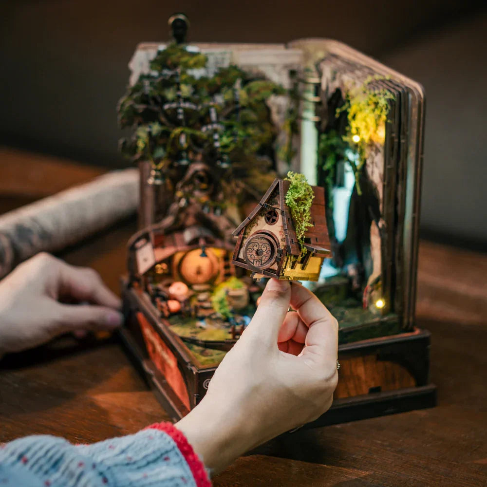 Eine Person hält ein Miniaturhaus mit Moos auf dem Dach über einem Diorama in einem Buch