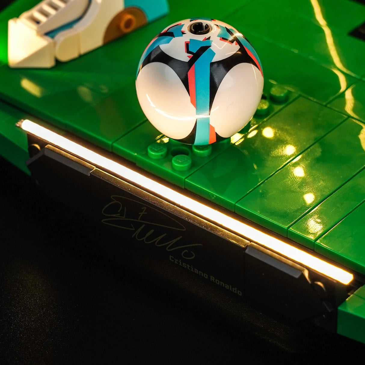 Detailaufnahme eines LEGO-Fußballs auf einem grünen Spielfeld, beleuchtet durch eine LED-Lichtleiste an der Kante, die eine schwarze Fliese mit Cristiano Ronaldos Unterschrift anstrahlt.