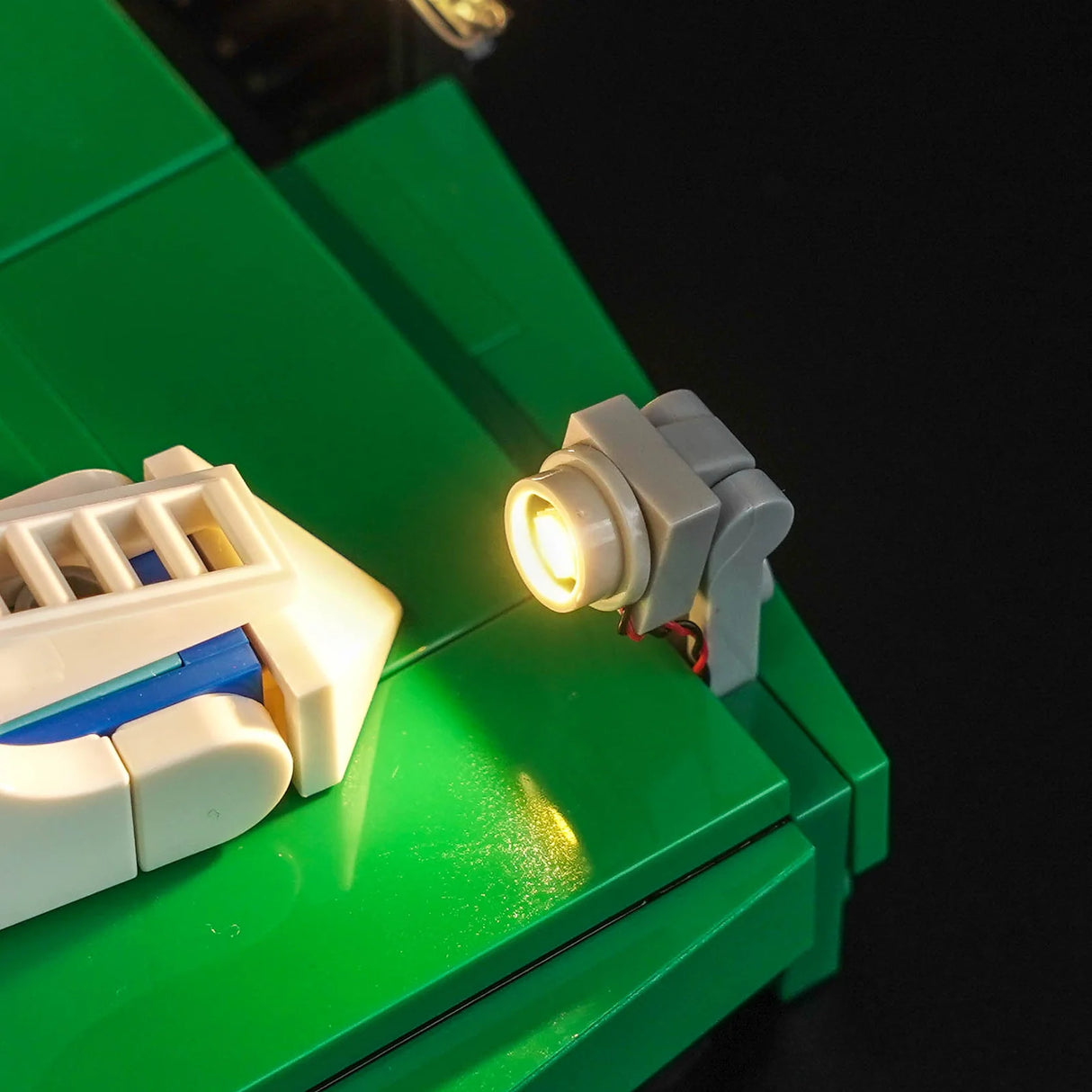 Nahaufnahme eines kleinen grauen LEGO-Scheinwerfers mit warmweißem LED-Licht aus dem BrickBling Beleuchtungs-Kit, montiert auf einer grünen LEGO-Platte neben einem weißen Fußballschuh.
