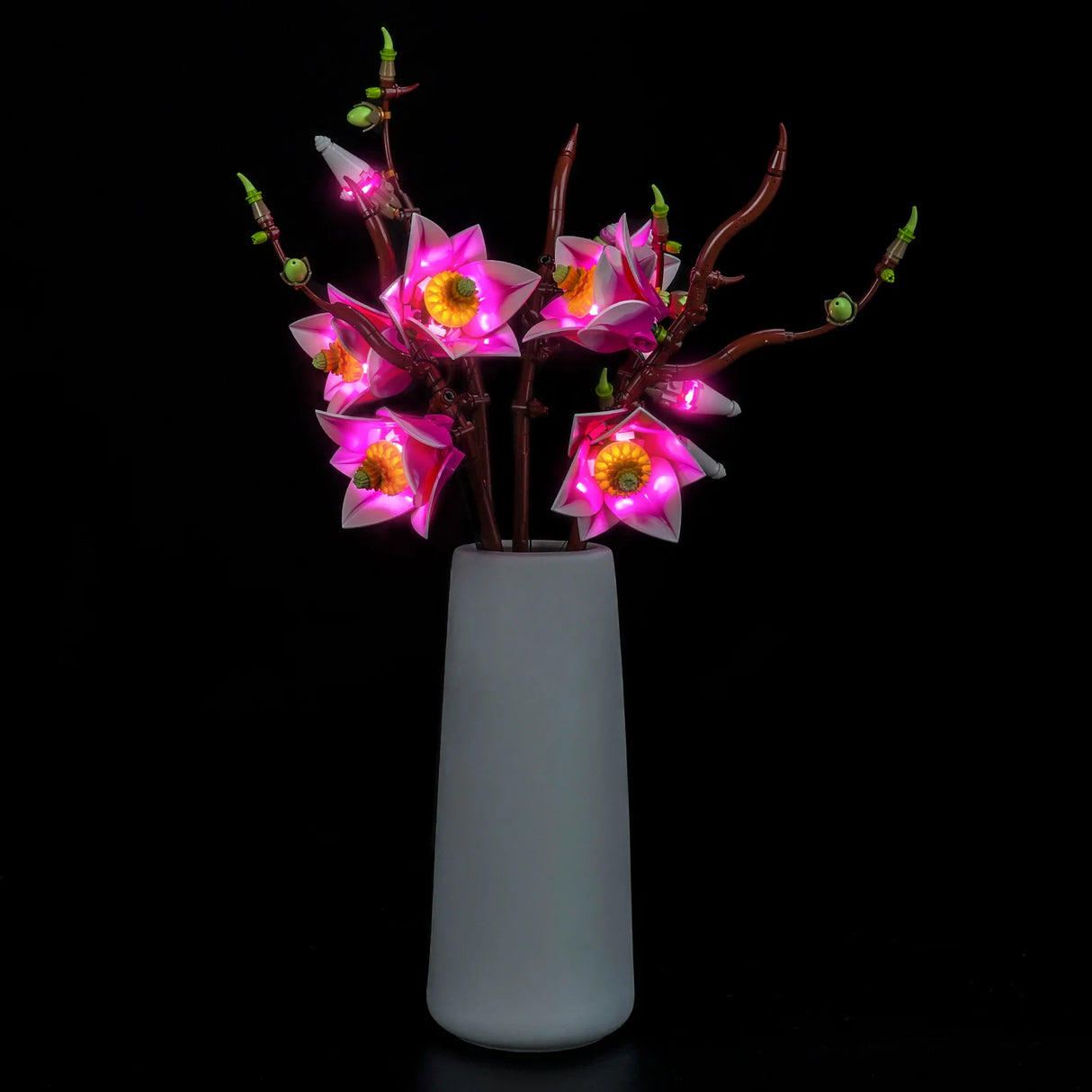Frontale Gesamtansicht der LEGO Magnolienzweige in einer grauen Vase mit eingeschalteter LED-Beleuchtung von BrickBling vor schwarzem Hintergrund.