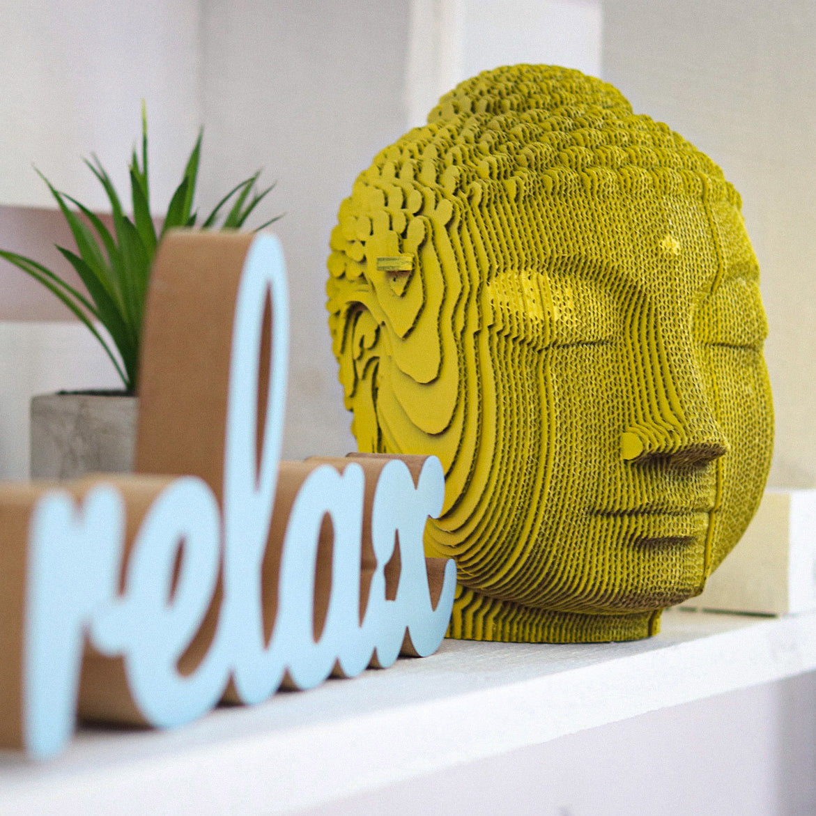Gelber Buddha Kopf aus Wellpappe neben einem Schriftzug Relax auf einem Regal
