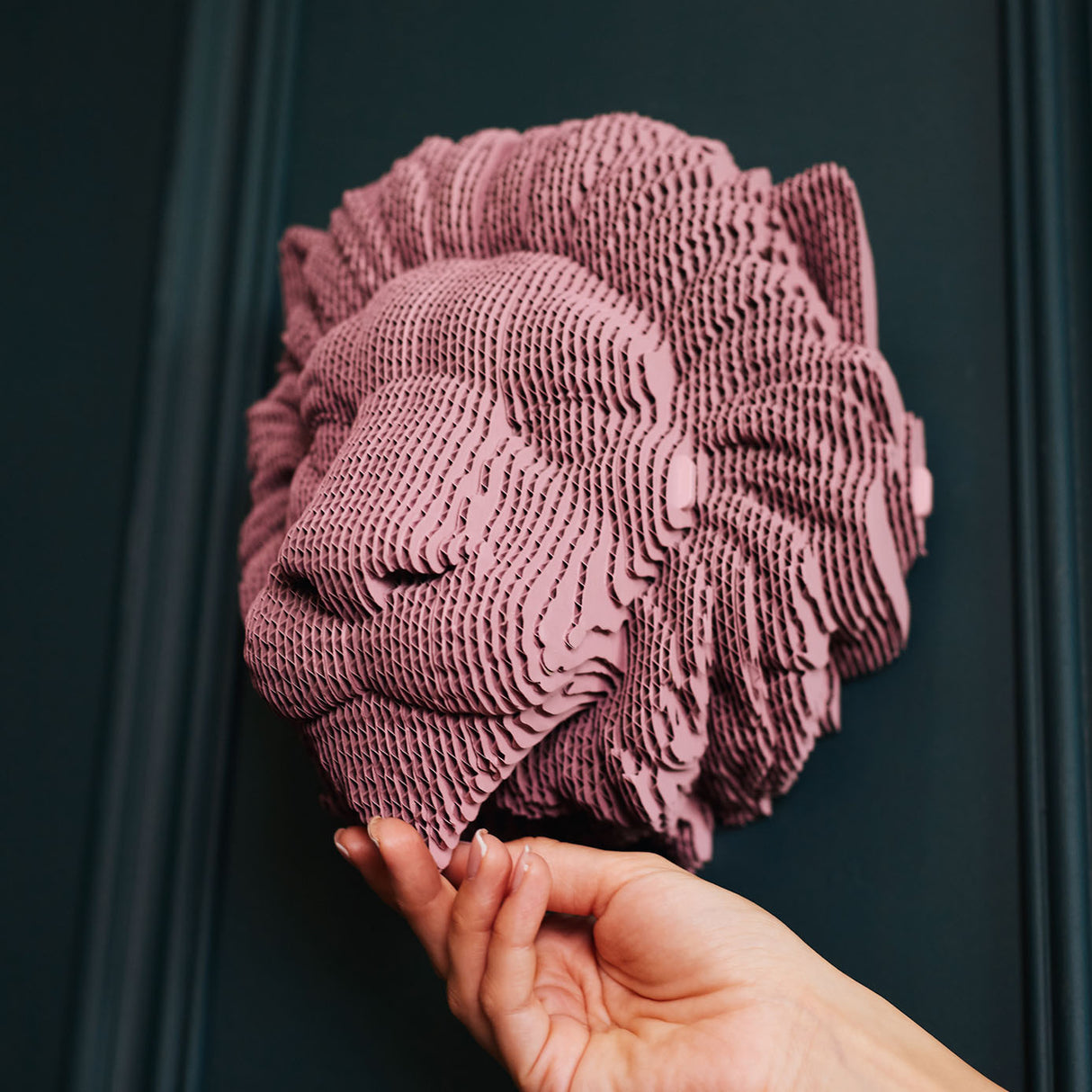 Hand hält eine rosa Löwenkopf Skulptur aus geschichtetem Karton vor einer dunklen Wand