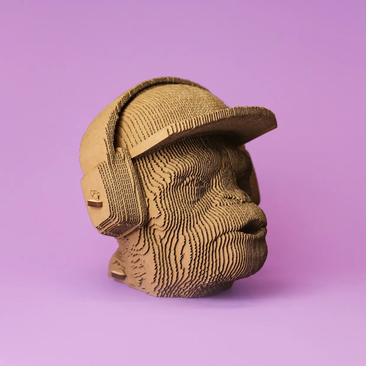 Skulptur eines Affenkopfes aus geschichtetem Karton mit Kopfhörern und Kappe vor lila Hintergrund