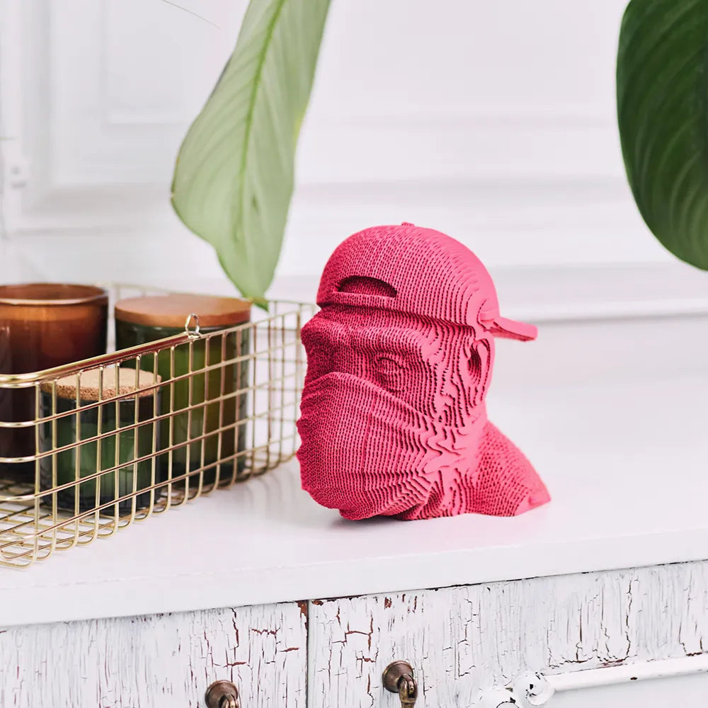 Pinke 3D gedruckte Skulptur eines Kopfes mit Kappe und Mundschutz auf einem weißen Tisch