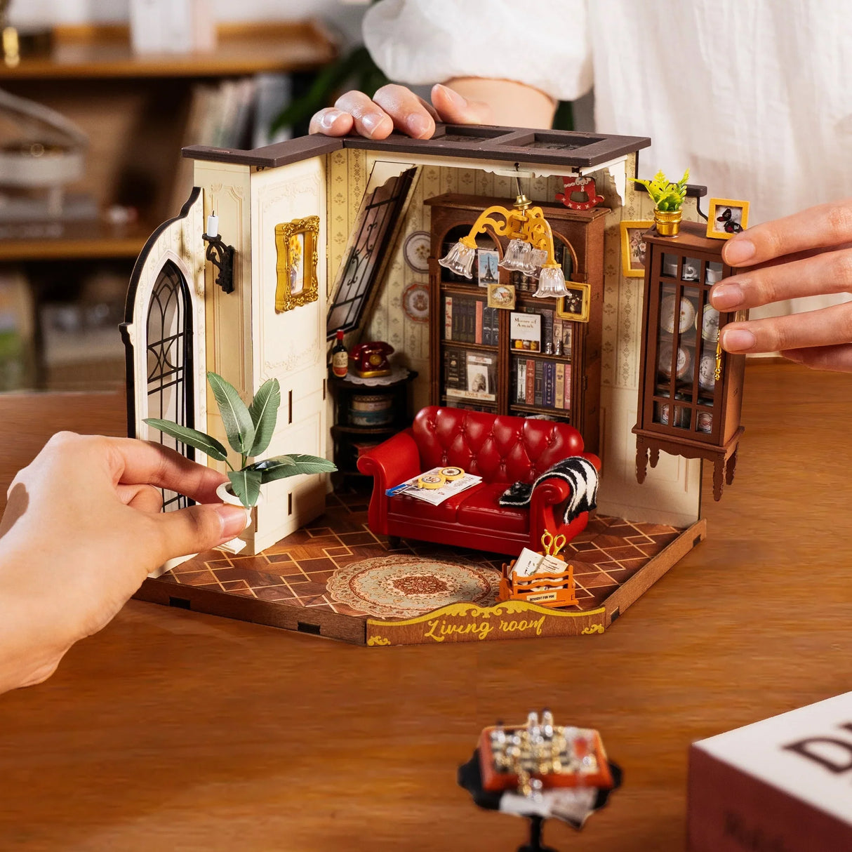Zwei Hände arrangieren Miniaturmöbel in einem Puppenhaus-Wohnzimmer mit rotem Sofa und Bücherregal