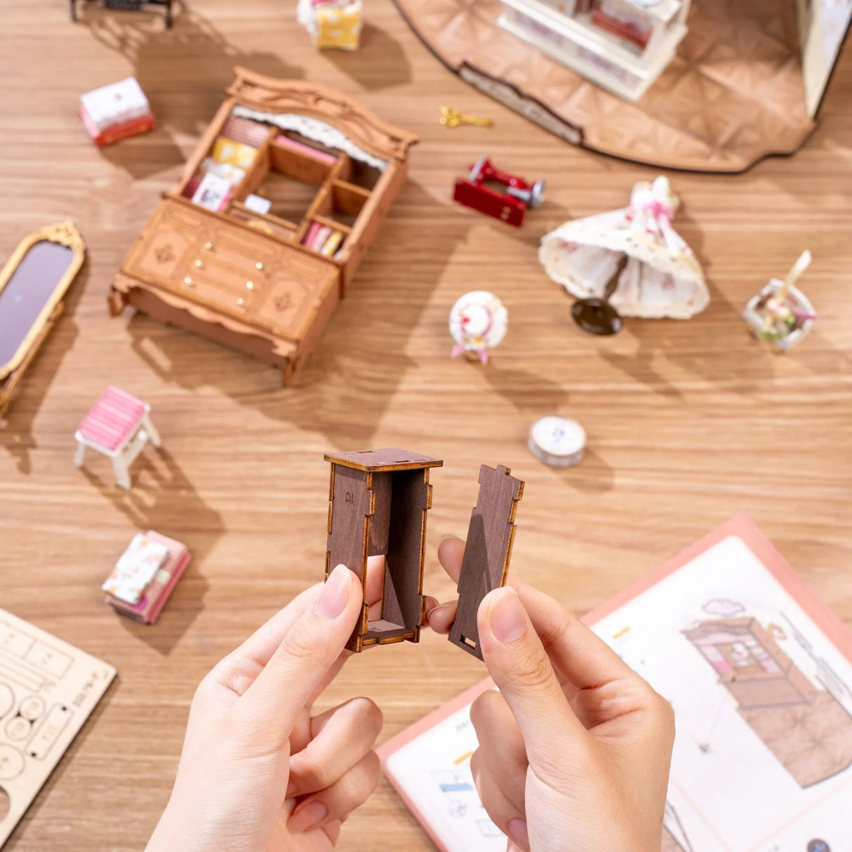 Hände halten zwei Holzteile eines Miniaturmöbels über einem Holztisch mit weiteren Miniaturmöbeln