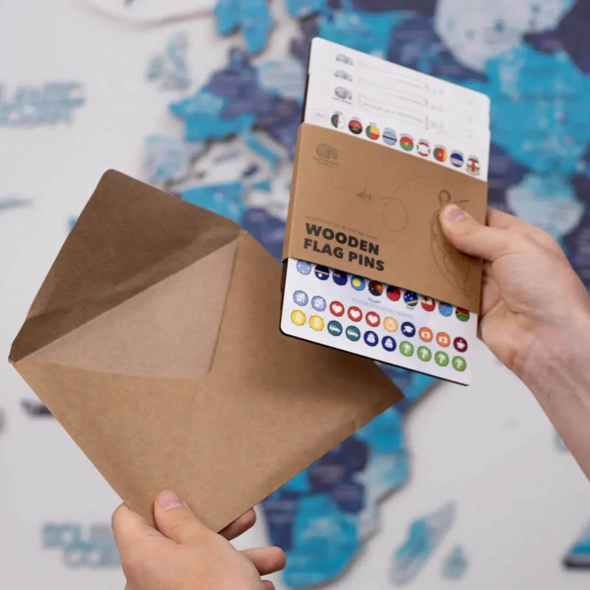 Hände halten einen braunen Umschlag und ein Set mit hölzernen Flaggenpins vor einer Weltkarte