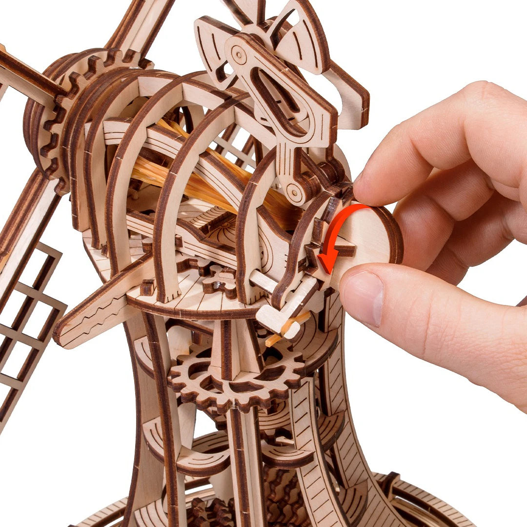 Hand dreht ein Zahnrad an einem hölzernen Windmühlenmodell mit sichtbaren Zahnrädern und Mechanismus