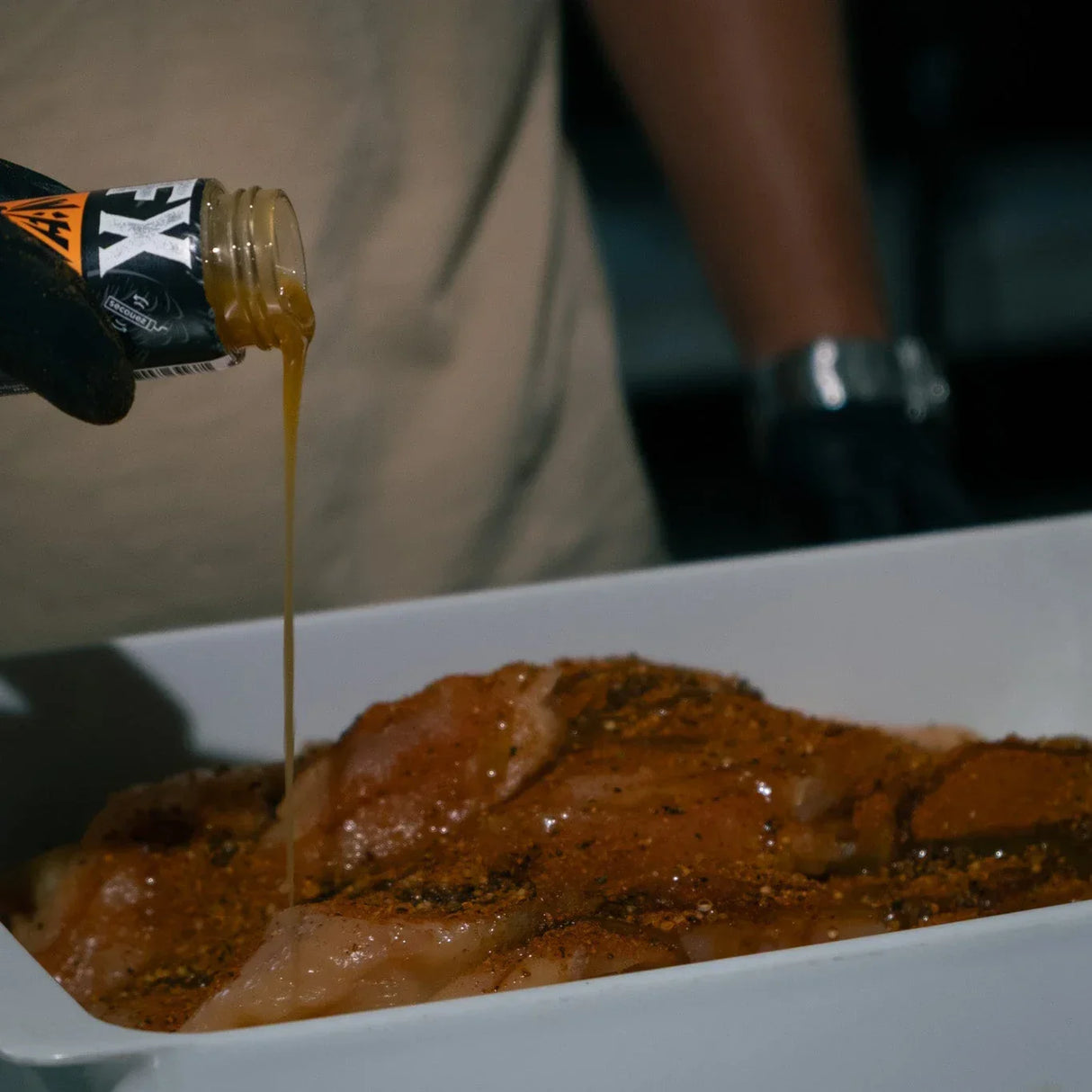 Eine Hand gießt Soße aus einer Flasche über gewürztes Hähnchen in einer weißen Schale