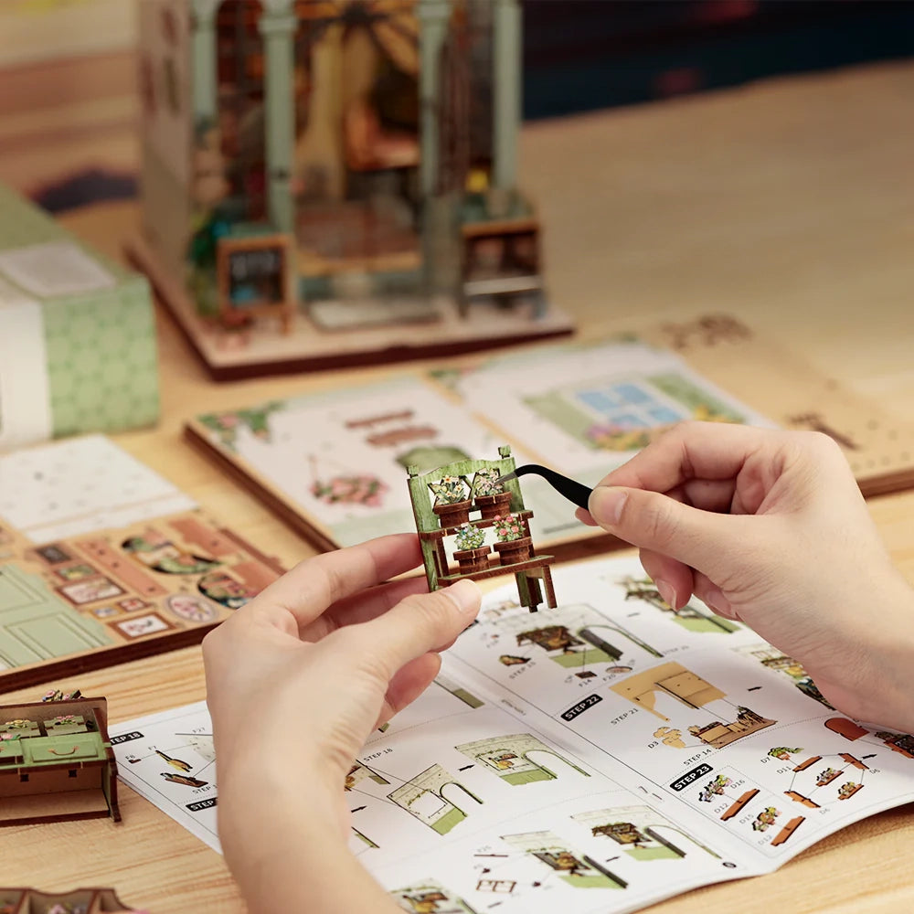 Hände bauen ein Miniatur-Pflanzenregal mit Pinzette, Anleitungsheft und fertigem Puppenhaus im Hintergrund