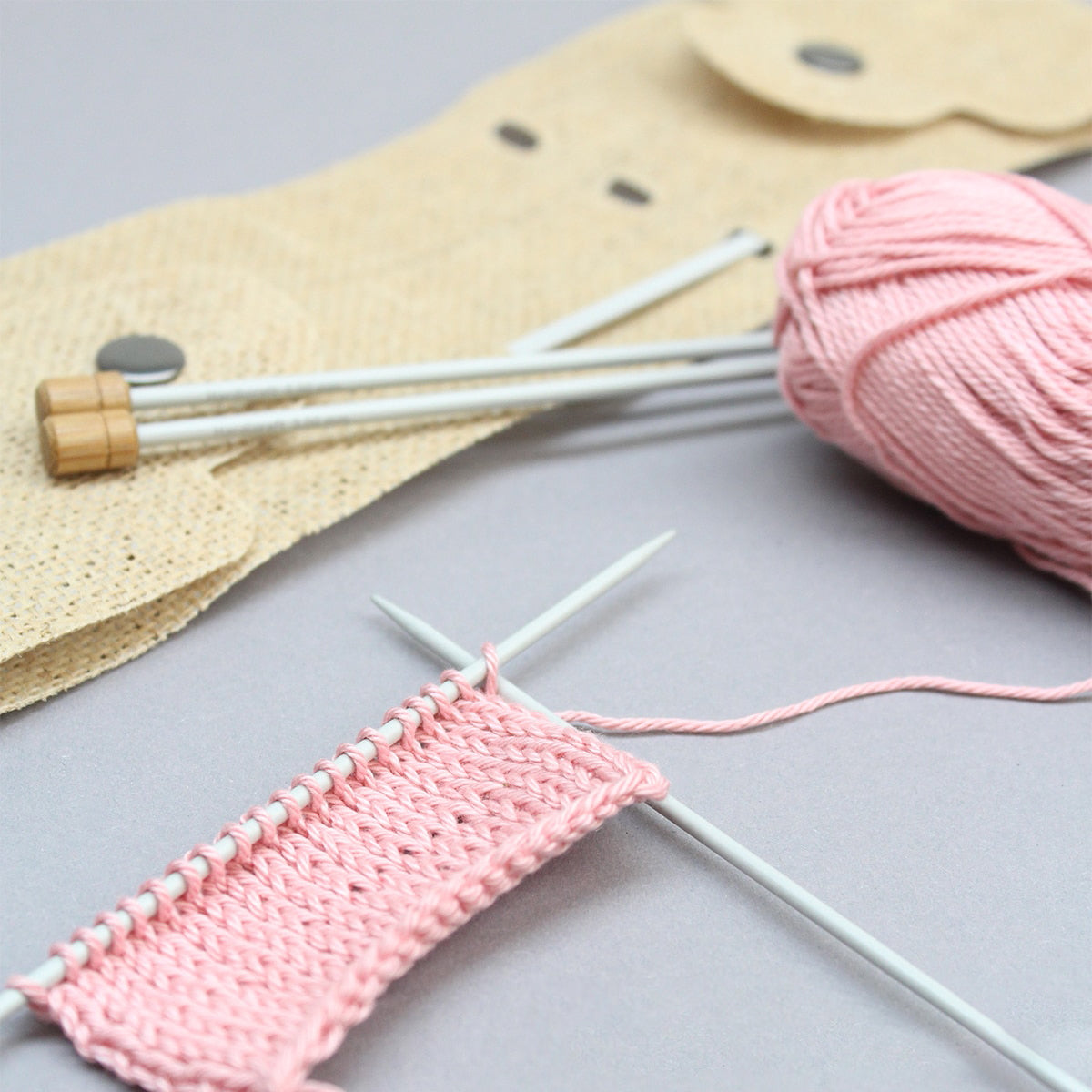 Rosa Garnknäuel mit Stricknadeln und einem beigen Etui auf grauem Untergrund
