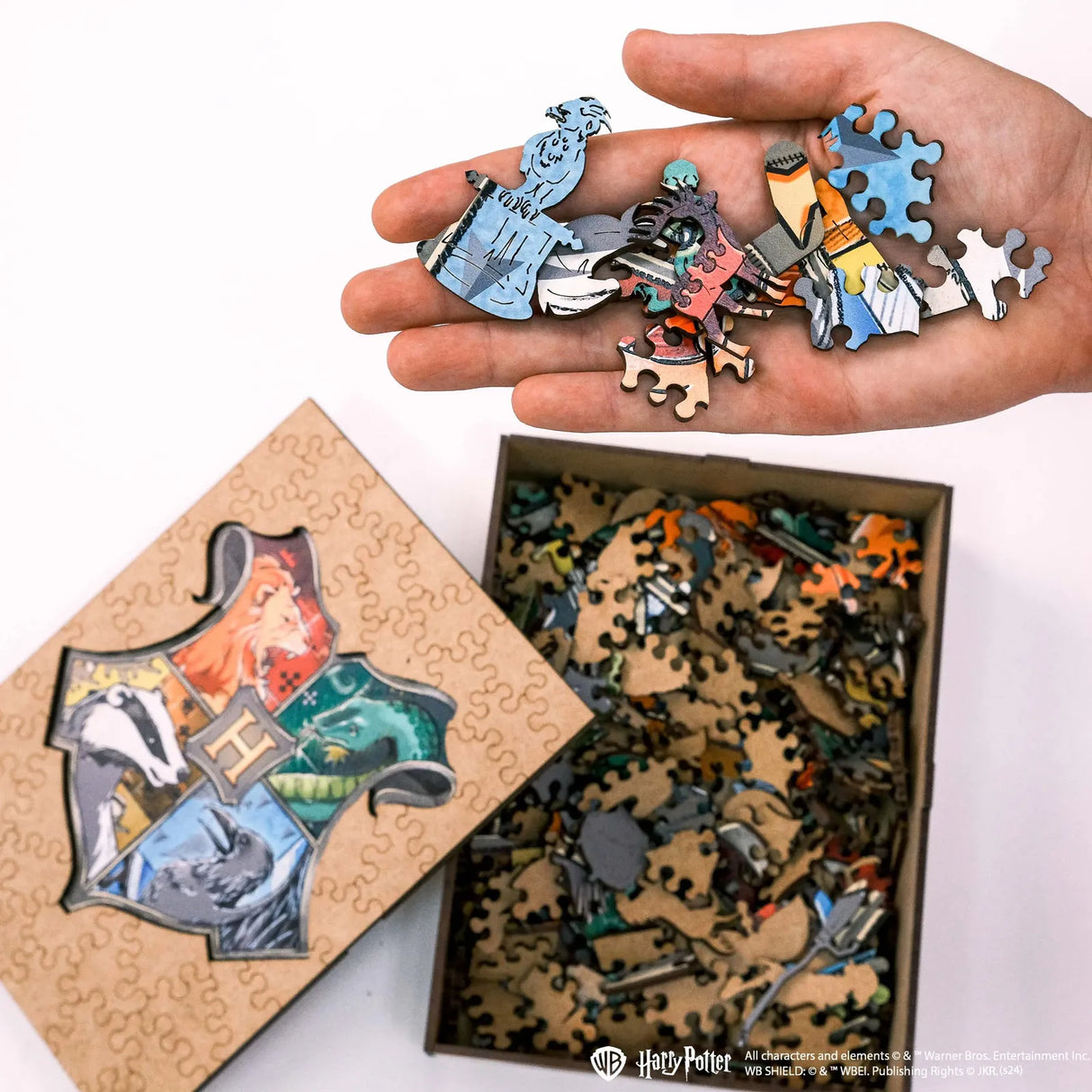Hand hält bunte Puzzleteile über einer Schachtel mit Harry Potter Wappen und weiteren Teilen
