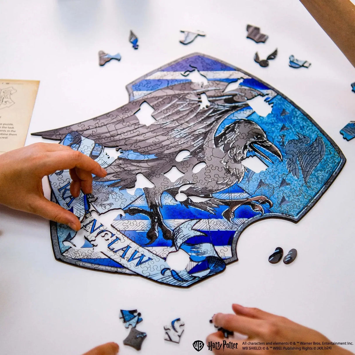 Hände legen ein Ravenclaw-Wappen-Puzzle mit einem Raben und blauen Streifen
