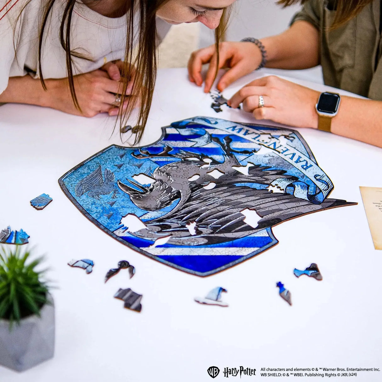 Zwei Personen legen ein Harry Potter Ravenclaw Wappen Puzzle auf einem weißen Tisch