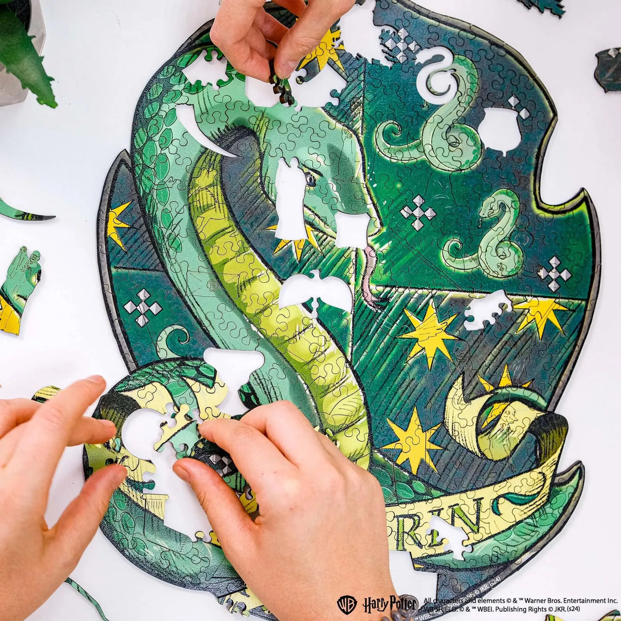 Zwei Hände setzen ein grünes Schlangenkopf-Puzzleteil in ein unvollständiges Slytherin-Wappenpuzzle