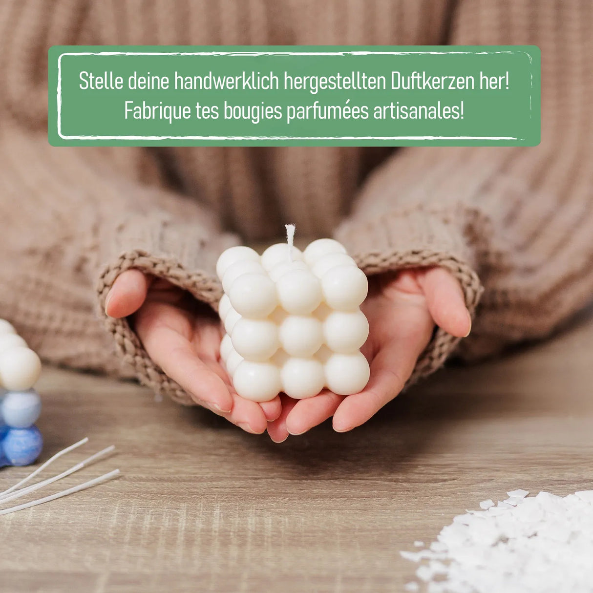 Hände halten eine weiße Blasenkerze über einem Holztisch mit Kerzenwachs und Dochten