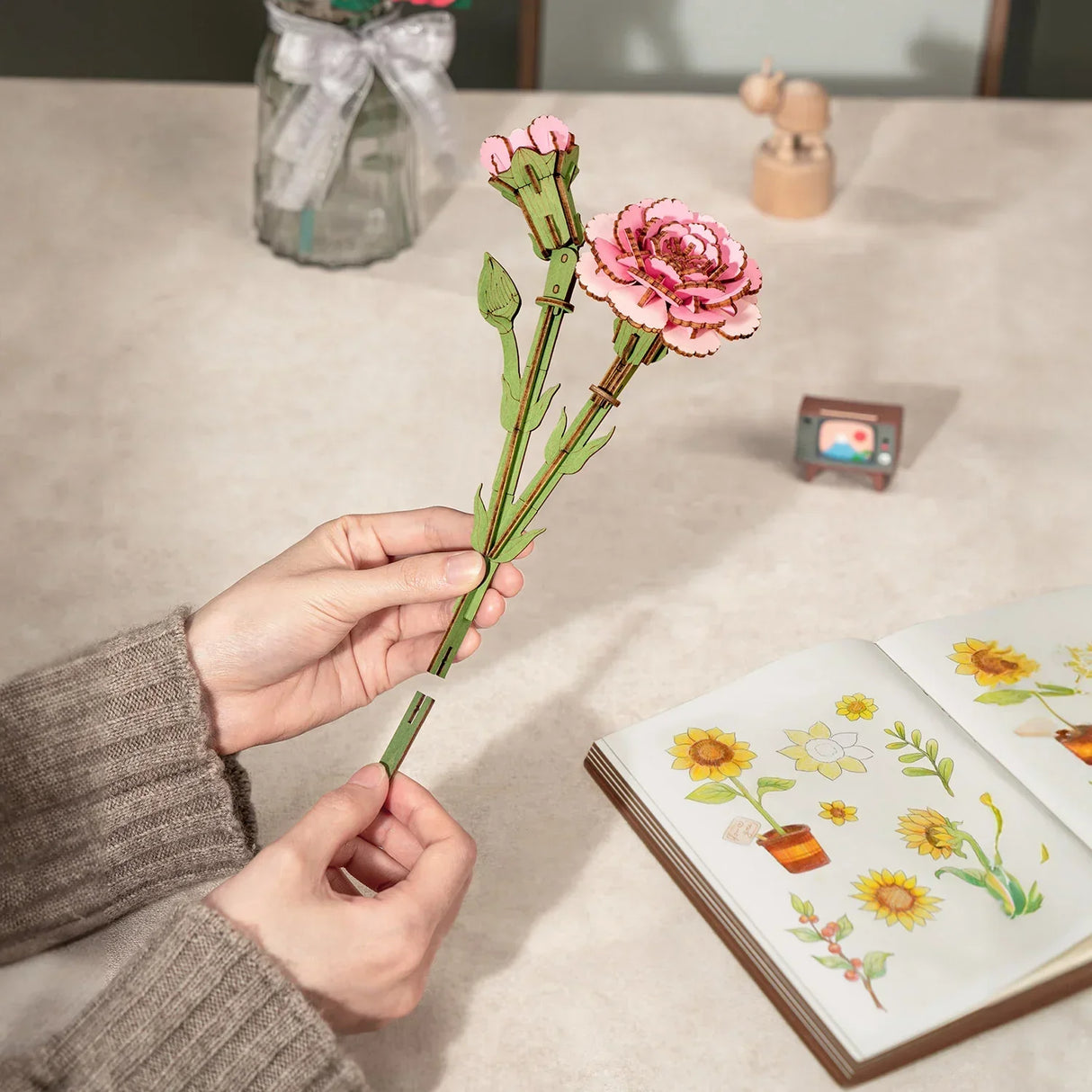 Hände halten eine zusammengesetzte Holzblume über einem offenen Notizbuch mit Sonnenblumenzeichnungen