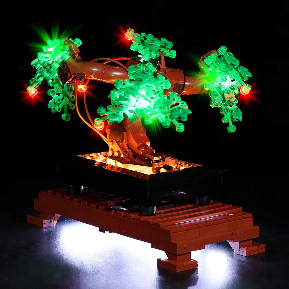 Rückansicht oder seitliche Ansicht des LEGO Bonsai-Baums, die die Verzweigung der Äste zeigt. Ein dünnes Kabel des LED-Lichtsets verläuft unauffällig entlang des Stammes. Die drei Hauptblattbereiche werden von hellen grünen und roten LEDs illuminiert, während die Unterseite des Podests taghell erleuchtet wird.
