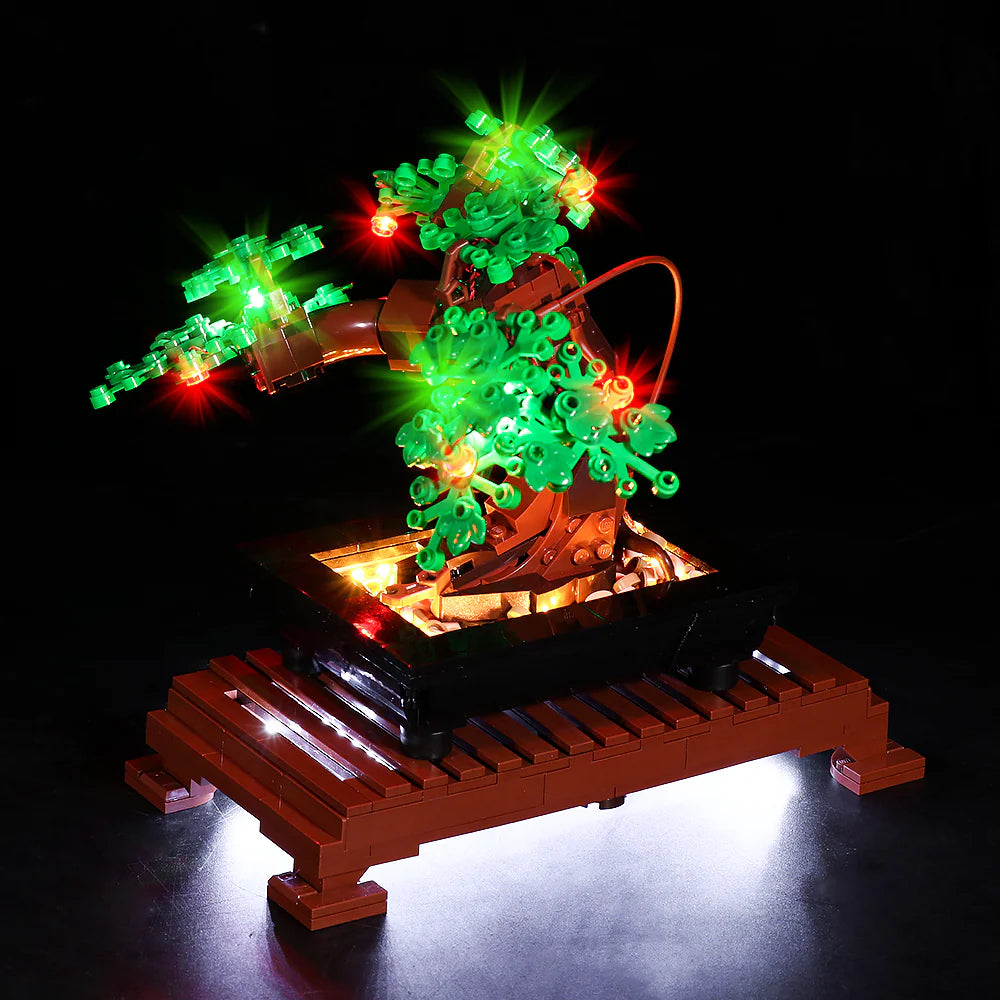 Detailansicht des beleuchteten LEGO Bonsai-Baums (Set 10281) von vorne. Der Fokus liegt auf dem Zusammenspiel der roten und grünen LED-Lichter im Laub und der warmweißen Ausleuchtung der kleinen Steine innerhalb der schwarzen Pflanzschale.