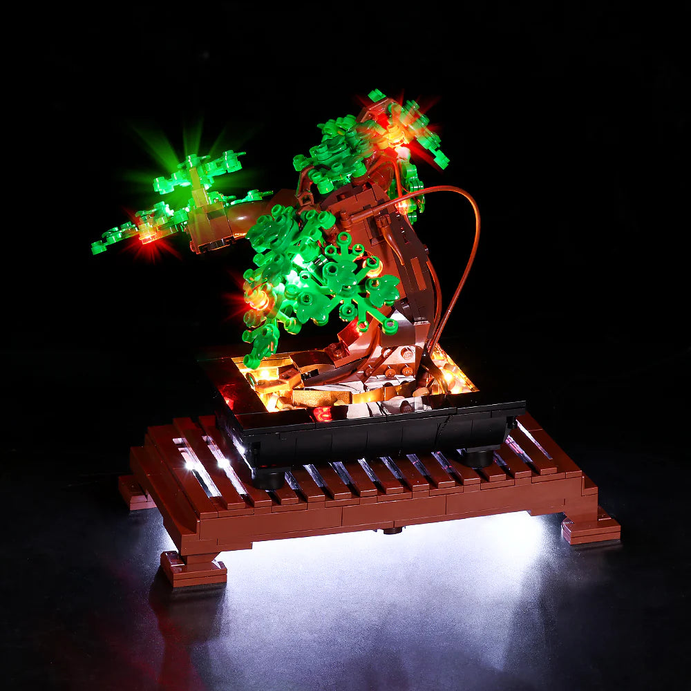 Perspektivische Schrägansicht von vorne links auf den LEGO Bonsai-Baum mit grünem Laub. Die LED-Beleuchtung setzt die Blätterkronen mit roten und grünen Lichtern in Szene. Warmes Licht strahlt aus der Schale nach oben und wirft einen hellen Schein durch das braune Podest nach unten auf den Boden.