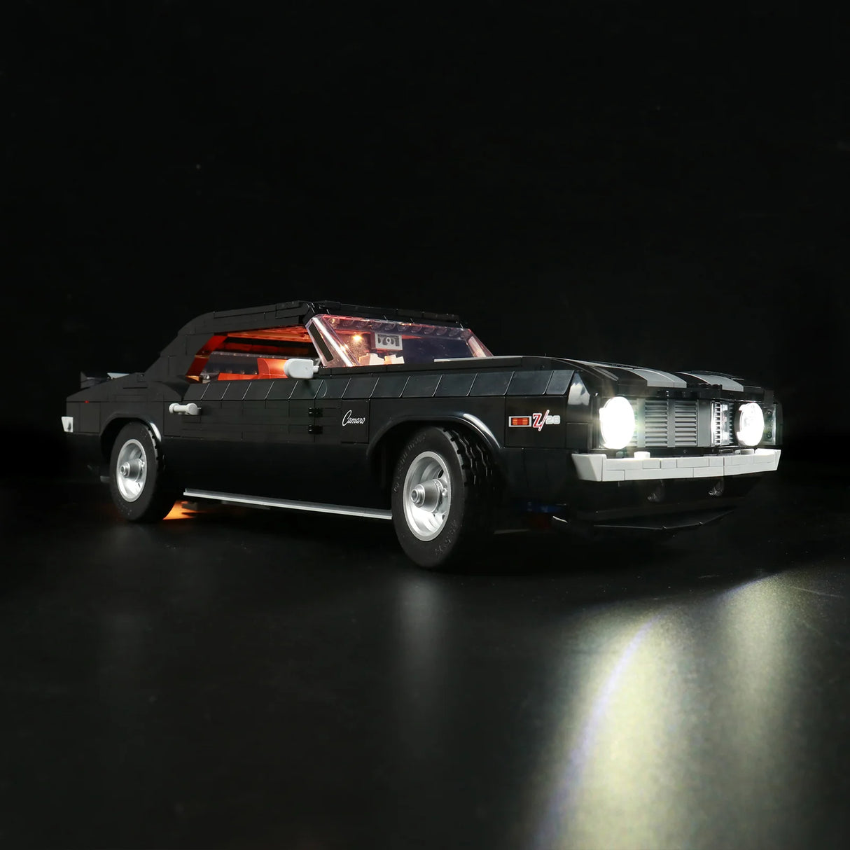 Schräge Frontansicht des schwarzen LEGO Chevrolet Camaro Z28 Modells mit LED-Licht-Upgrade. Die hellen Scheinwerfer werfen einen Lichtkegel auf den Boden, der Innenraum leuchtet rot.