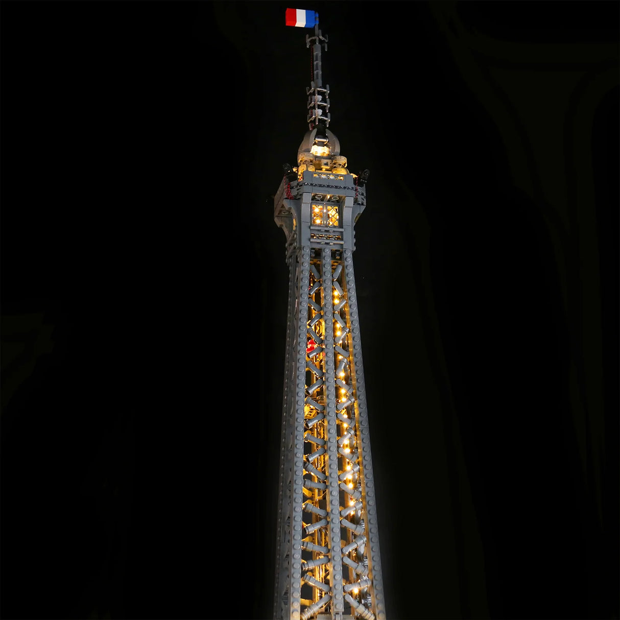 Nahaufnahme der beleuchteten Spitze des LEGO Eiffelturms 10307 mit warmer LED-Beleuchtung und französischer Flagge vor dunklem Hintergrund. 44883432702180