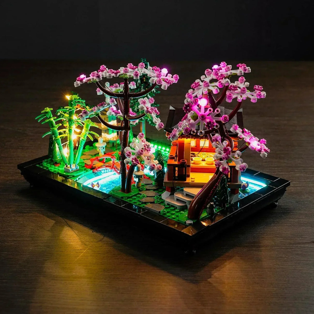 Schräge Draufsicht des LEGO 10315 Sets mit LED-Lichtern bei Nacht, die den asiatischen Garten, die Steinpfade und die rosa Kirschblüten stimmungsvoll hervorheben.