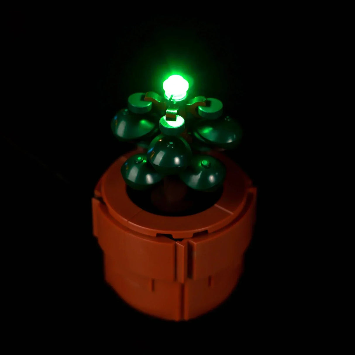Eine kleine grüne LEGO Pflanze im braunen Topf aus dem Set 10329, deren Spitze durch ein integriertes grünes LED-Licht hell erstrahlt.