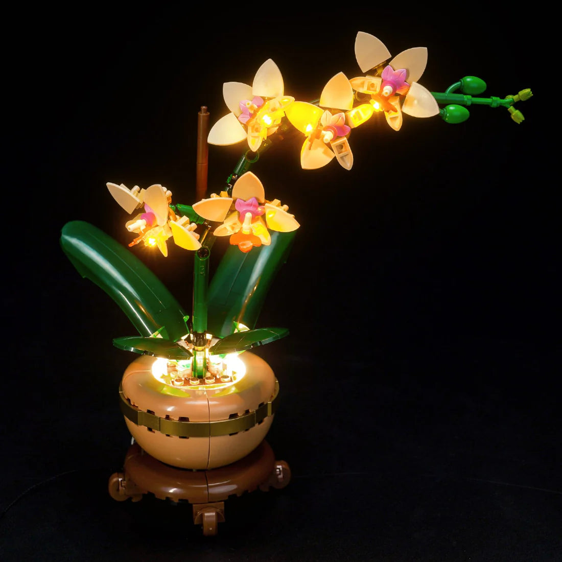 Beleuchtete LEGO Orchidee mit gelben Blüten und grünen Blättern in einem braunen Topf auf schwarzem Hintergrund