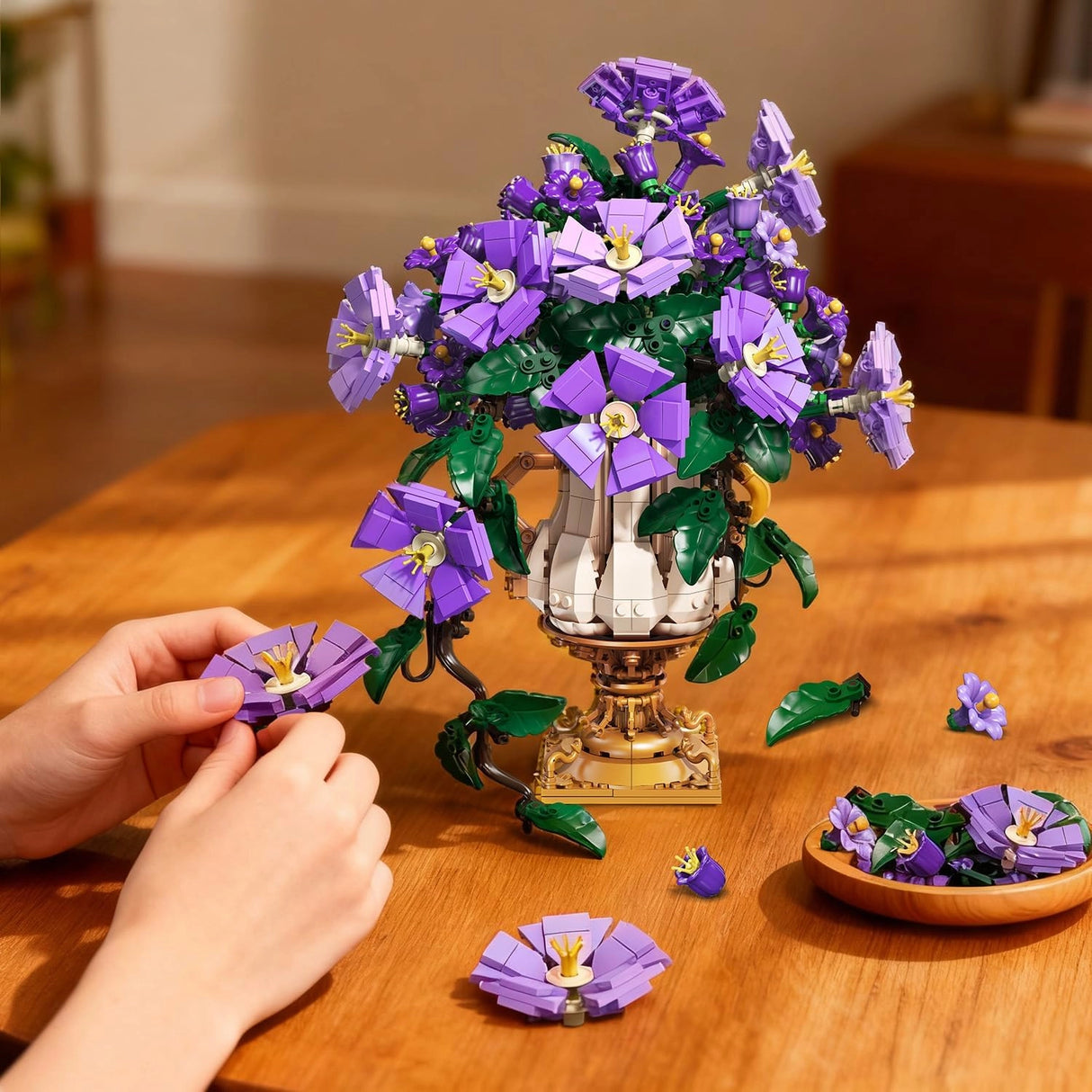 Hände bauen lila Lego-Blumen und Blätter in eine weiße Vase auf einem Holztisch