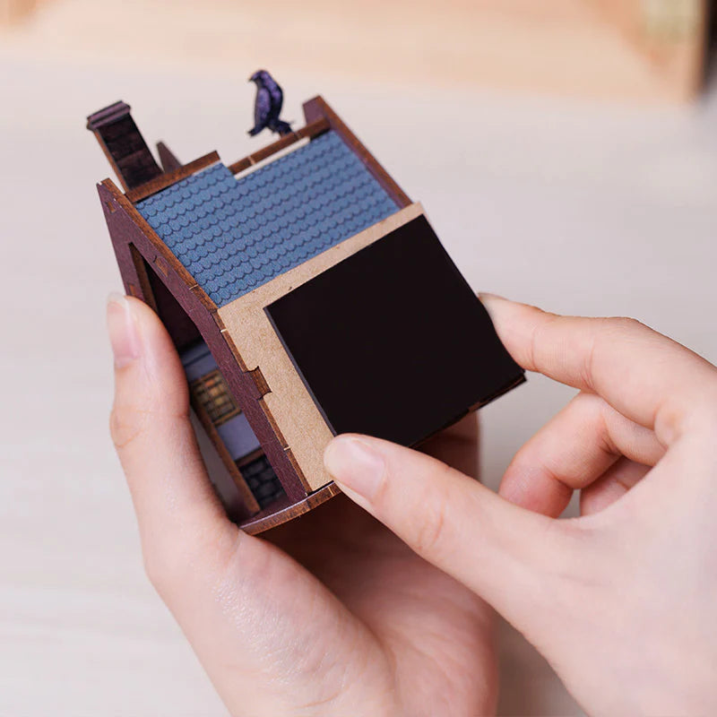 Hände halten ein Miniaturhaus mit blauem Dach und einem kleinen schwarzen Vogel auf dem Dachfirst