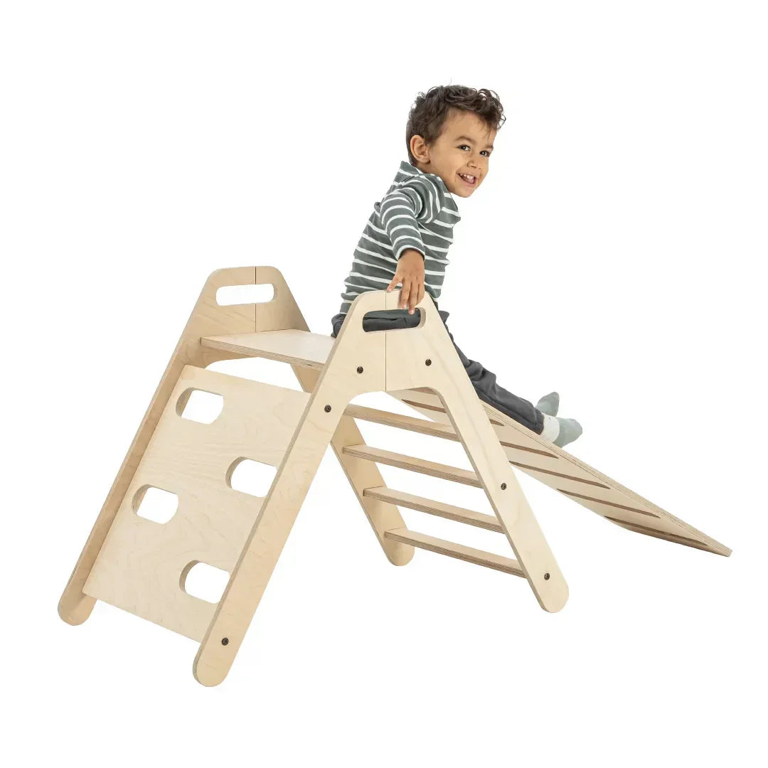 Ein lächelndes Kleinkind klettert auf das Mamatoyz Kletterdreieck aus Holz mit Rutschbrett-Aufsatz.