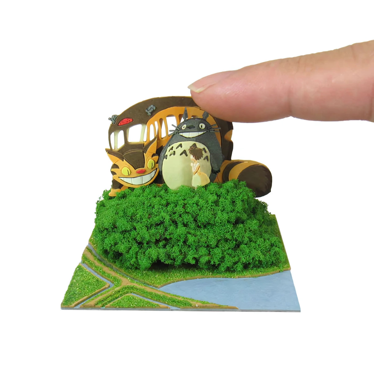 Finger berührt Totoro und Catbus Modell auf einer grünen Landschaft mit Fluss