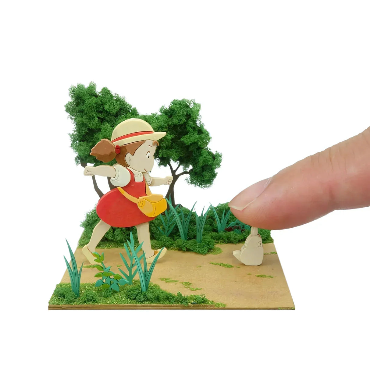 Miniatur-Diorama mit Mädchen in rotem Kleid und Strohhut, das auf einem Pfad geht, während ein Finger eine kleine weiße Figur berührt