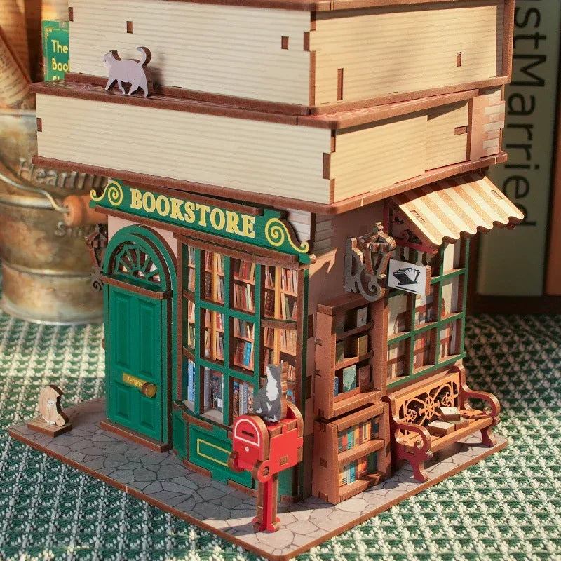 Miniatur-Buchhandlung mit grüner Tür und Fenster, darüber gestapelte Bücher, Katzenfiguren und ein roter Briefkasten