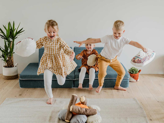 Drei Kinder spielen mit Kissen vor einem blauen Sofa und einem Lagerfeuer aus Kissen
