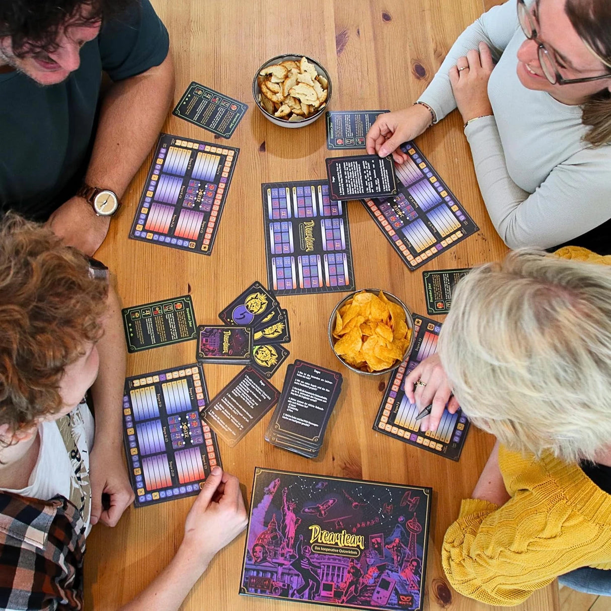 Vier Personen spielen das Brettspiel Dreamteam an einem Holztisch mit Spielkarten und Schalen voller Chips