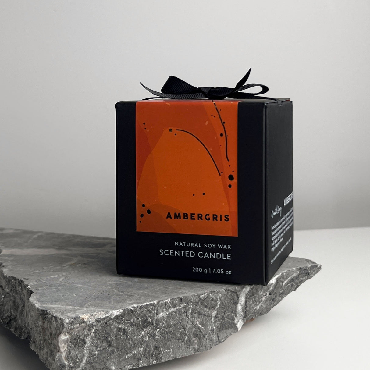 Schwarze Duftkerzenverpackung mit orangefarbenem Etikett auf einem grauen Stein