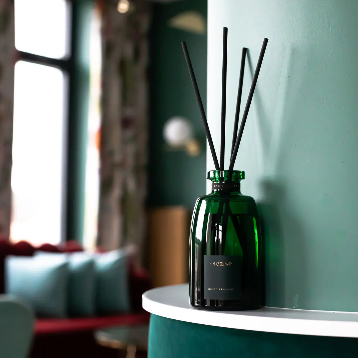 Grüner Glasdiffusor mit schwarzen Stäbchen auf einem weißen Regal vor einer grünen Wand