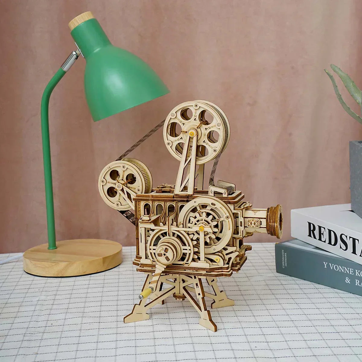Mechanisches Holzmodell eines Filmprojektors auf einem Tisch neben einer grünen Schreibtischlampe