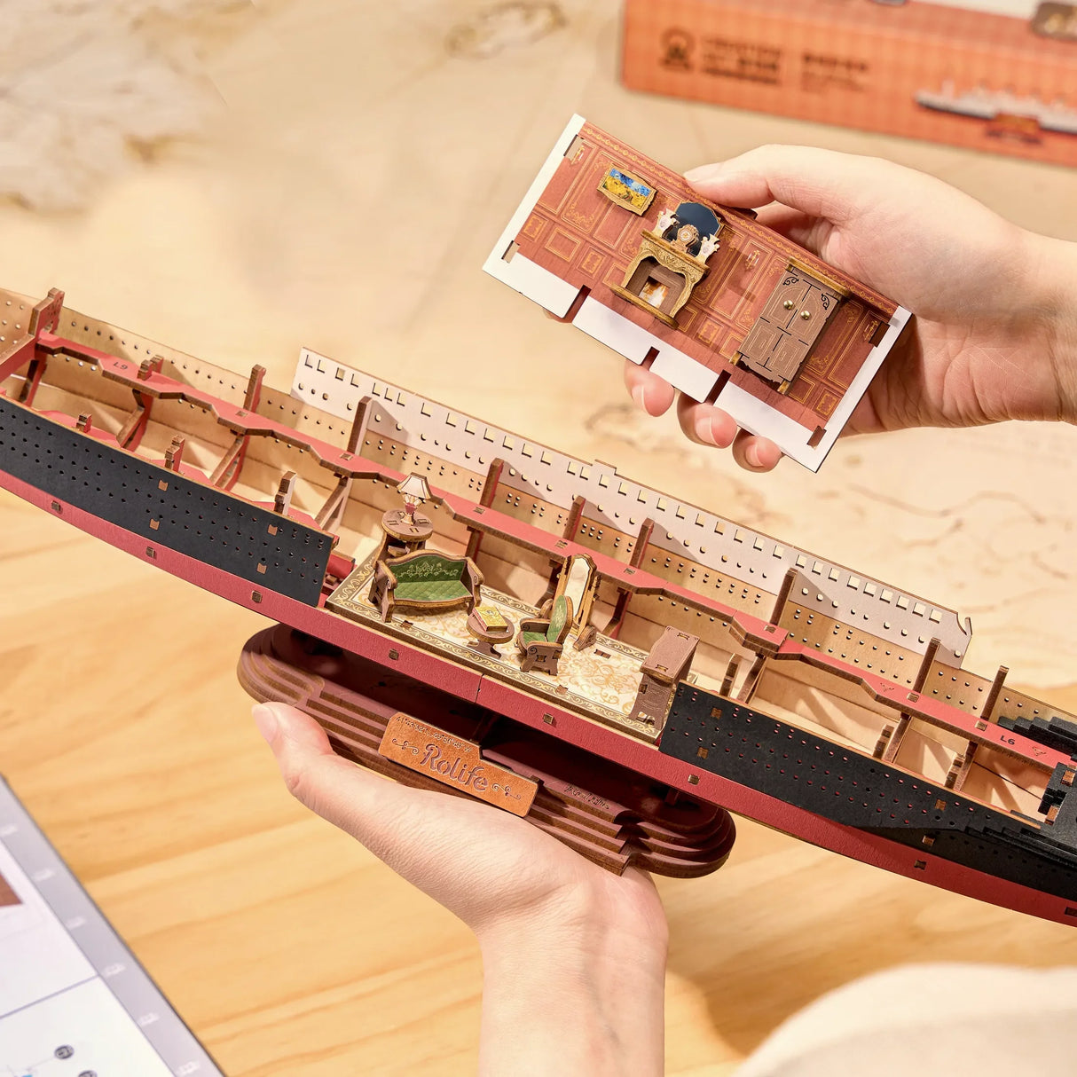 Hände halten ein Modellschiff mit sichtbaren Innenräumen und ein separates Wandstück mit Kamin
